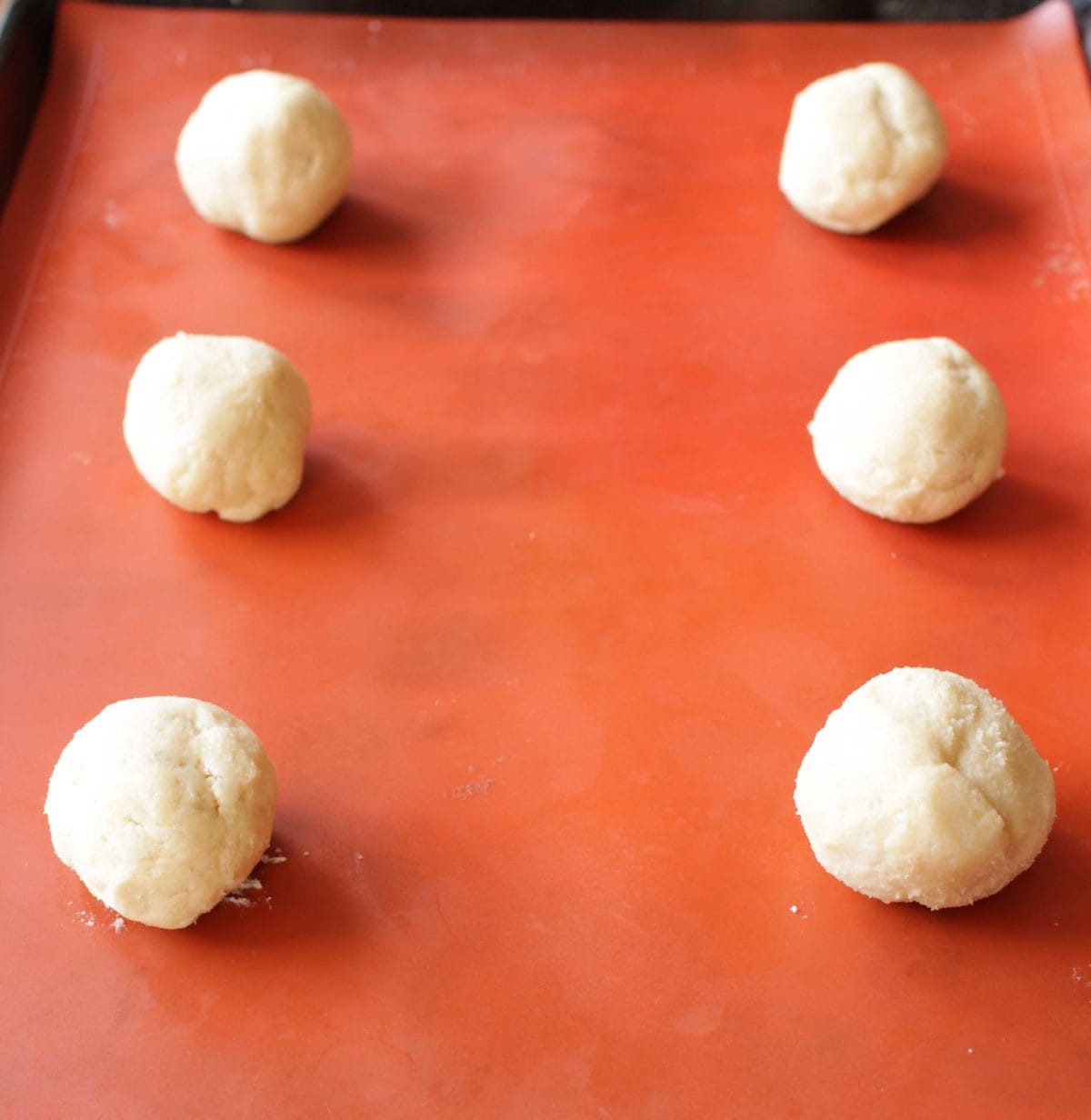 6 raw cookie dough balls on an orange silicone mat on a baking sheet.