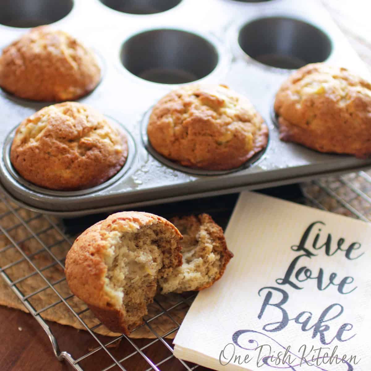 Four banana muffins in a muffin tin and a muffin split in half on a cooling rack.