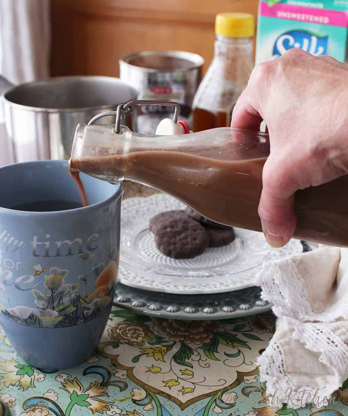 Pouring chocolate coffee creamer into a mug of coffee that is on a metal tray with a small plate of cookies.