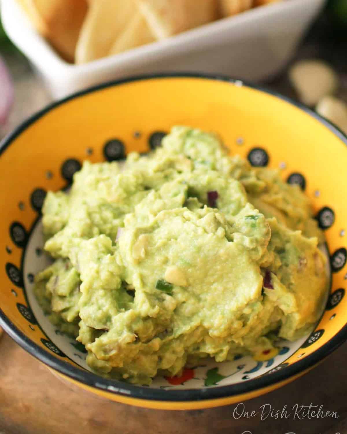 A small bowl of guacamole on a metal tray with a small bowl of homemade tortilla chips, an onion, garlic cloves, limes, and cilantro.