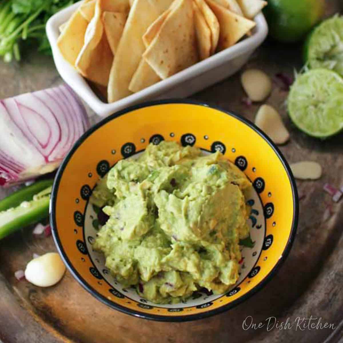a bowl of guacamole on a silver tray next to chips and onions and limes.