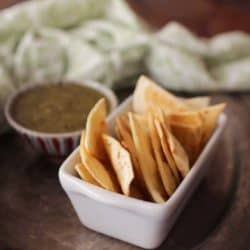 a white rectangular dish holding homemade tortilla chips next to a blue bowl filled with green salsa on a silver tray