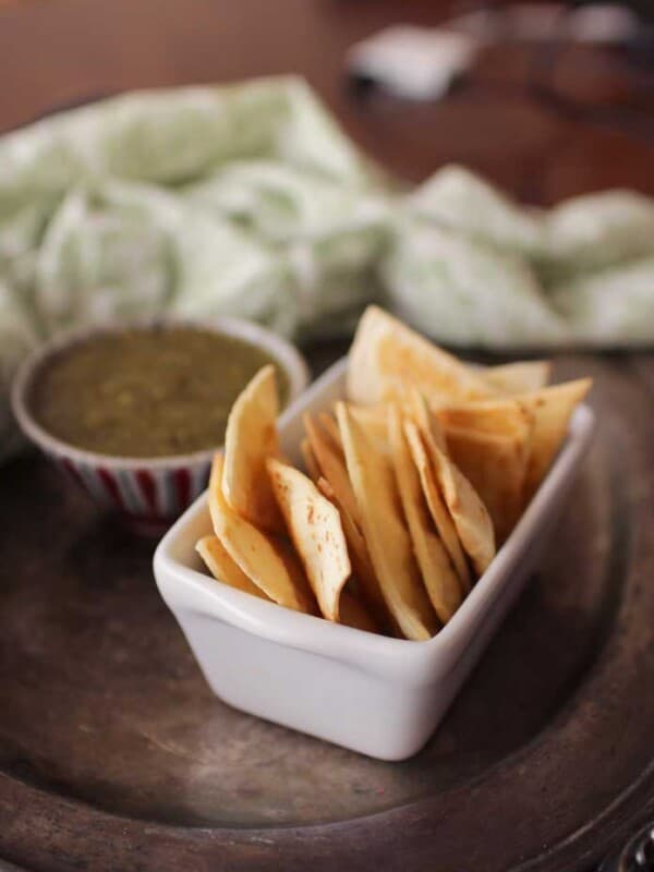 a white rectangular dish holding homemade tortilla chips next to a blue bowl filled with green salsa on a silver tray