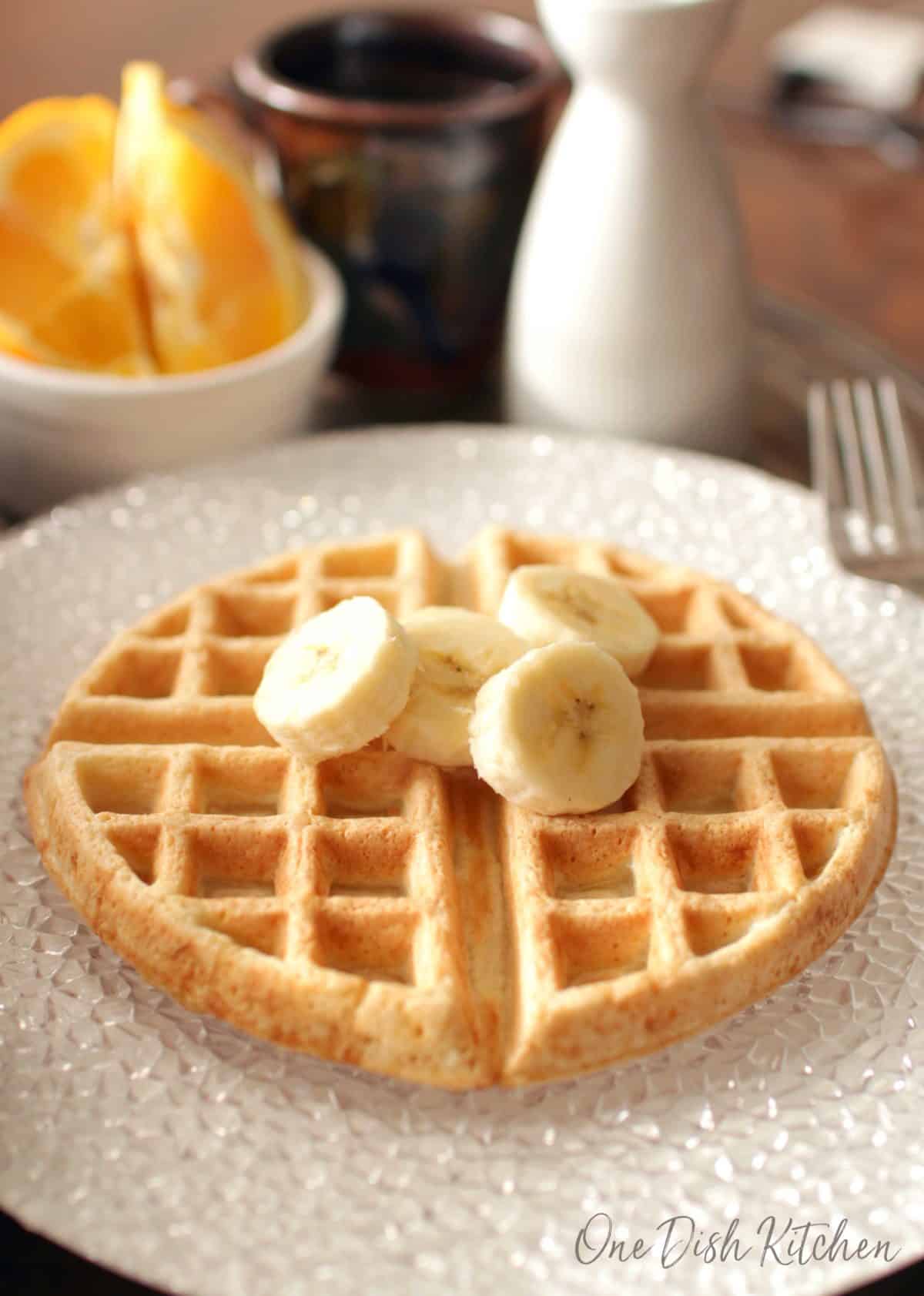 A waffle topped with banana slices on a large plate with a mug of coffee and orange slices in the background.