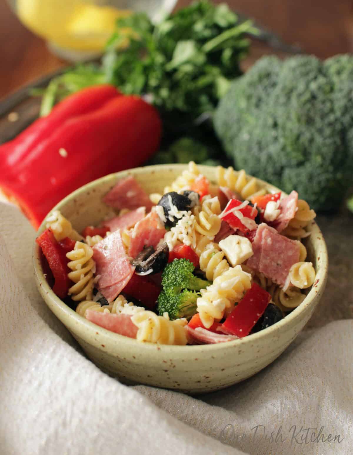 a bowl filled with italian pasta salad next to fresh broccoli on a silver tray.