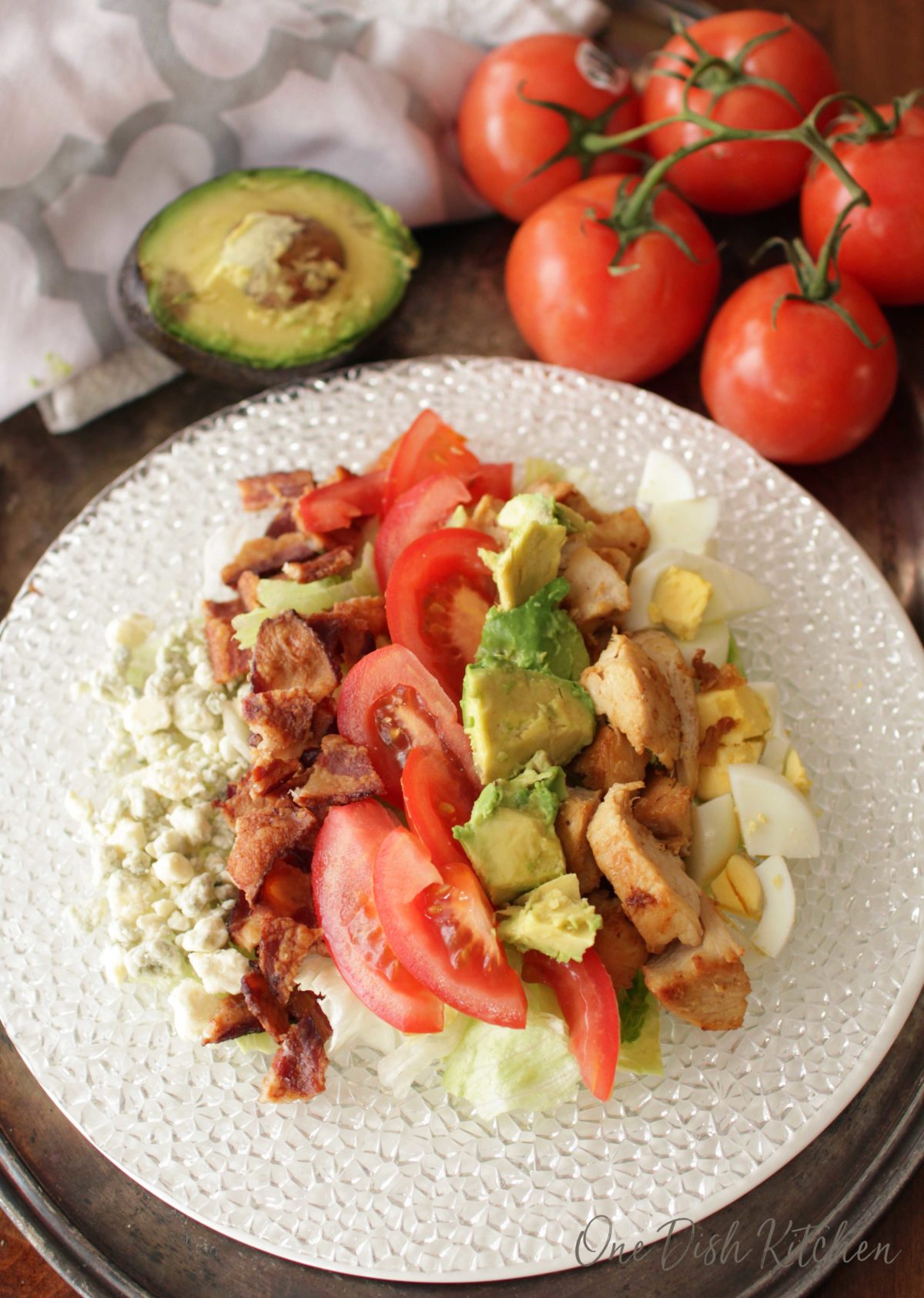 an overhead view of a cobb salad on a silver tray next to an avocado and 5 tomatoes
