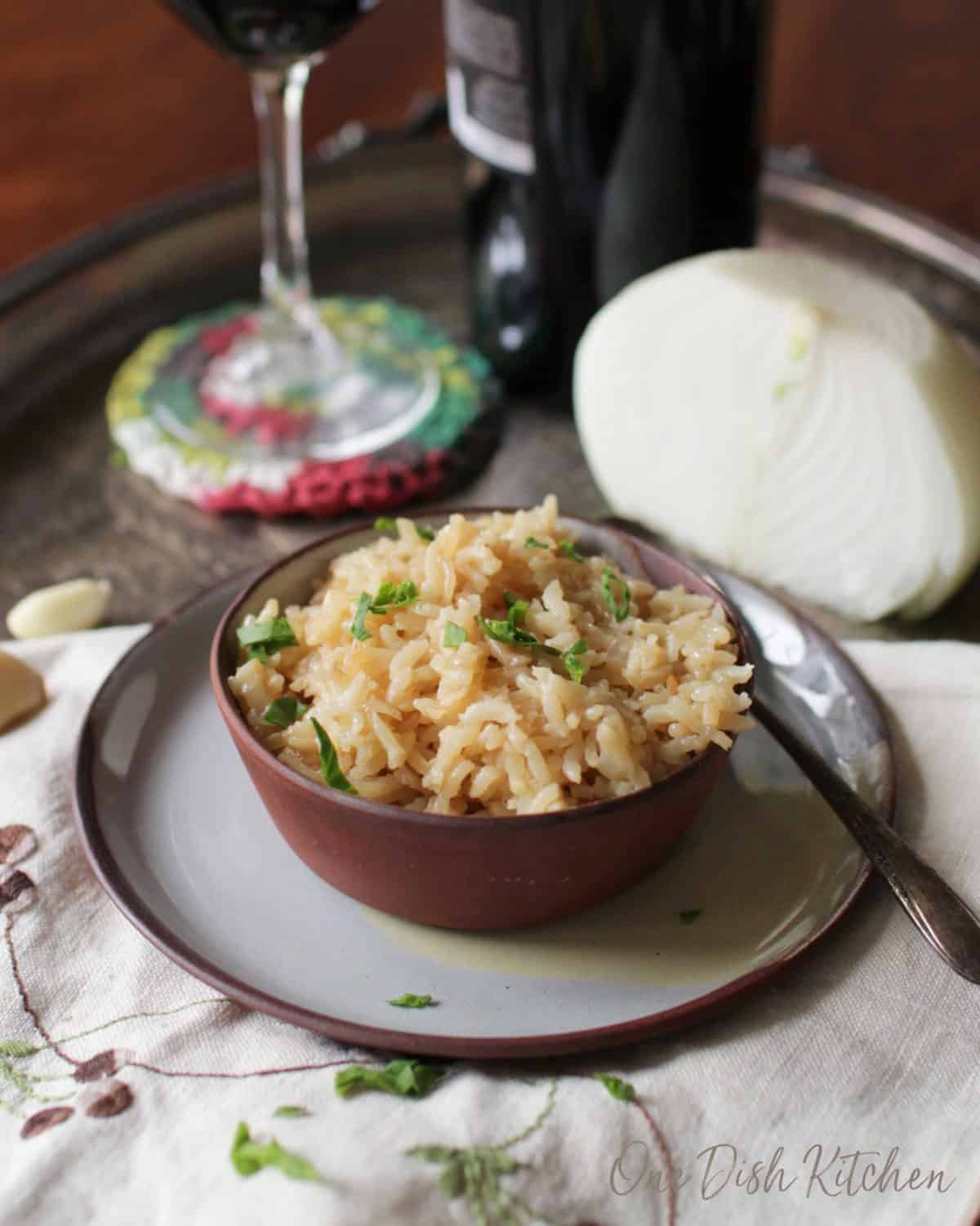 A bowl of rice pilaf plated on a metal tray next to half an onion, garlic cloves, and a glass of red wine 