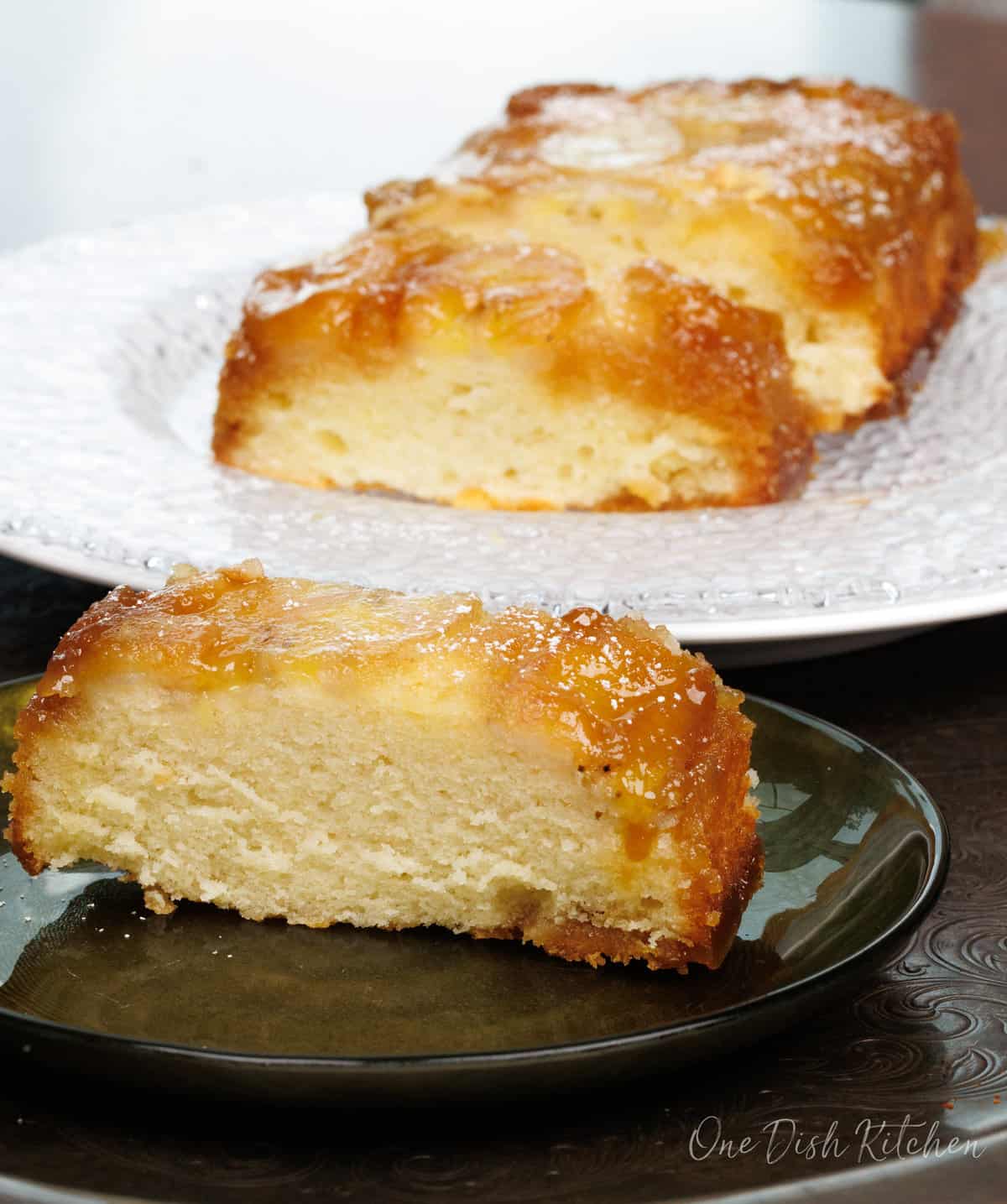 a slice of banana upside down cake on a green plate next to the cake loaf on another large plate.