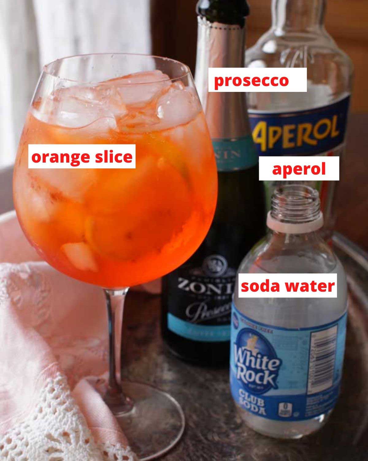 an aperol spritz on a brown table next to a bottle of prosecco, club soda and aperol