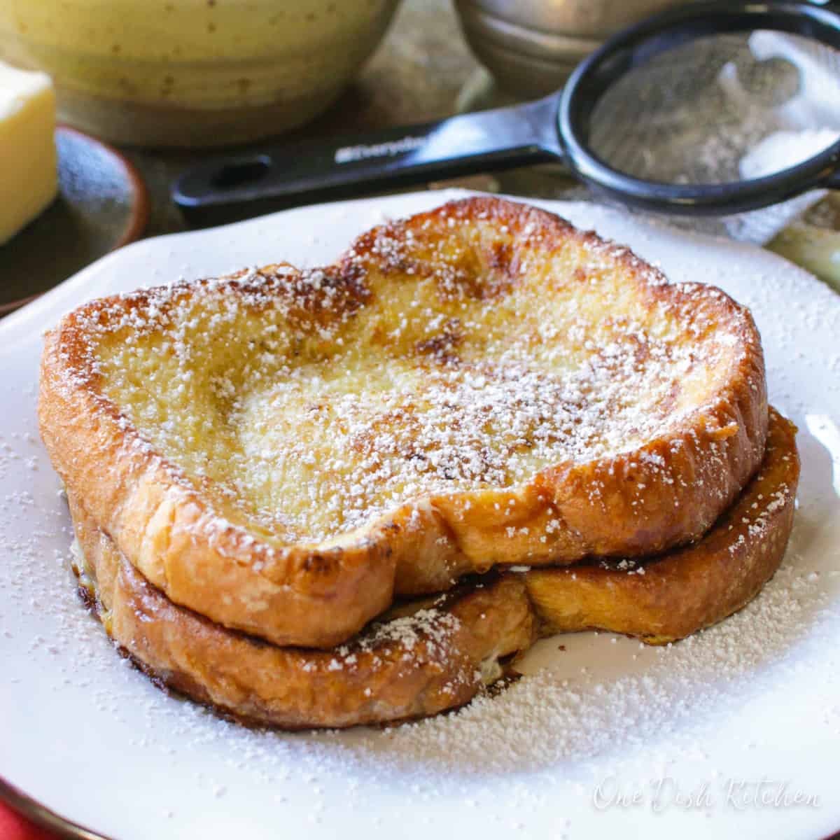 Two french toast slices dusted with powdered sugar on a metal tray with a stick of butter, a bowl of watermelon slices, and a metal container of maple syrup.