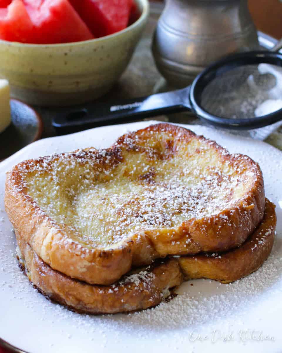 Two french toast slices dusted with powdered sugar on a metal tray with a stick of butter, a bowl of watermelon slices, and a metal container of maple syrup.