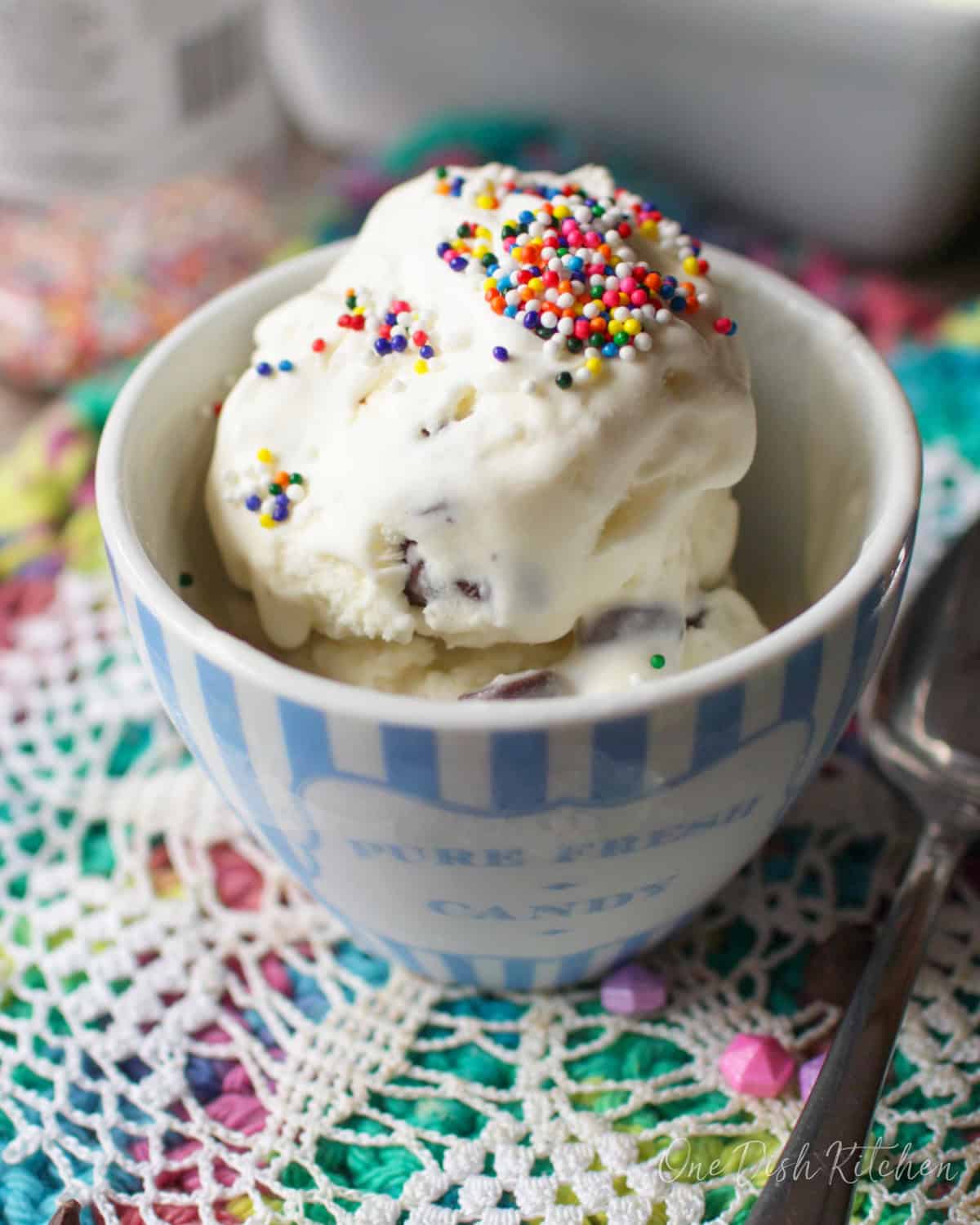 A small bowl of mint chocolate chip ice cream topped with rainbow sprinkles on a colorful surface next to a spoon and scattered chocolate chips.