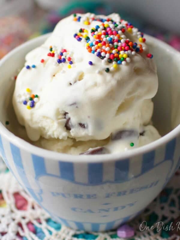 A small bowl of mint chocolate chip ice cream topped with rainbow sprinkles on a colorful surface next to a spoon and scattered chocolate chips.