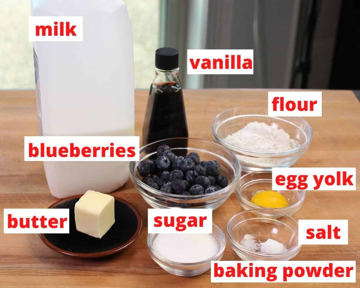 ingredients in blueberry muffins including flour, egg, sugar, and blueberries on a brown table.
