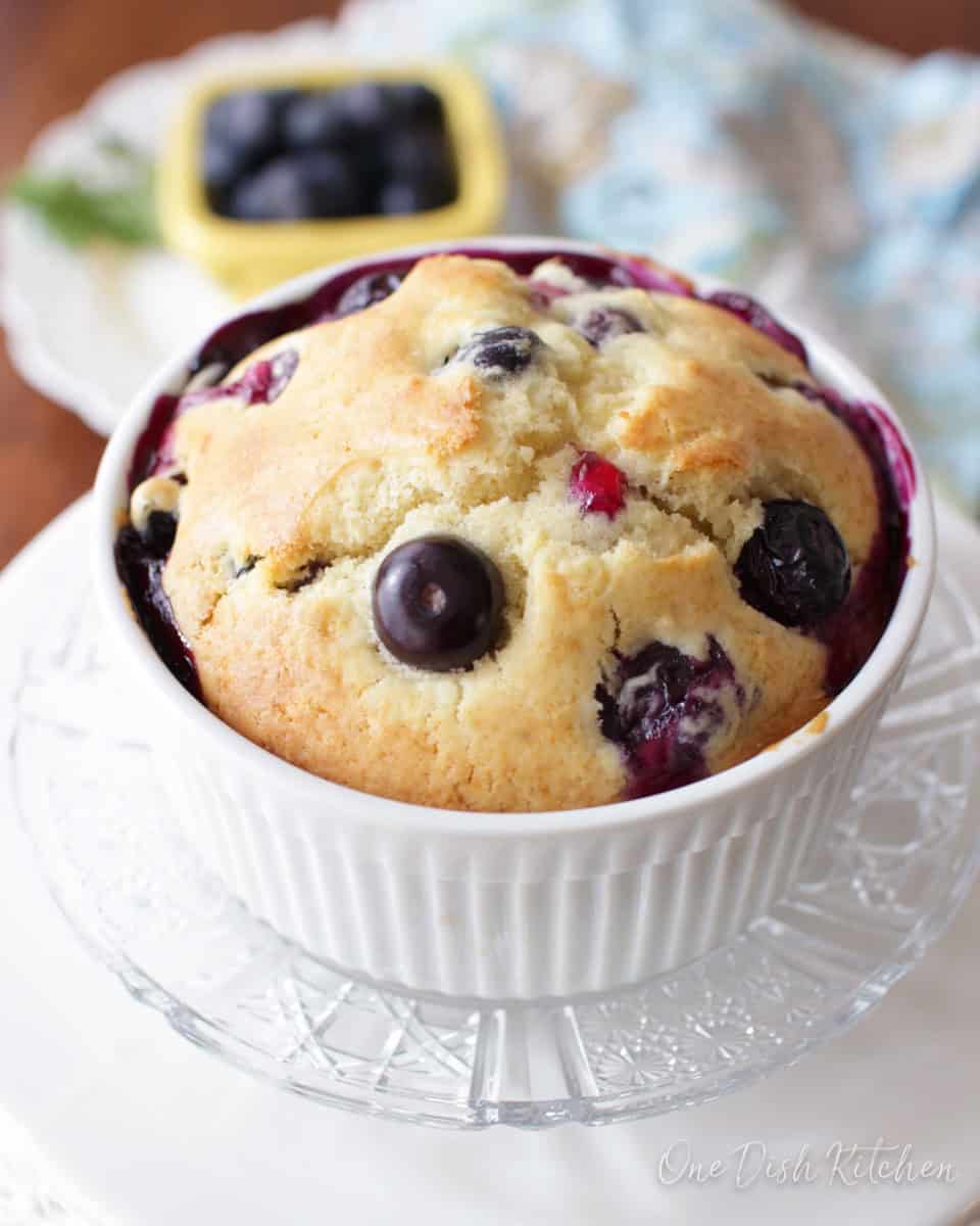 A blueberry muffin in a ramekin plated next to a small bowl of blueberries.