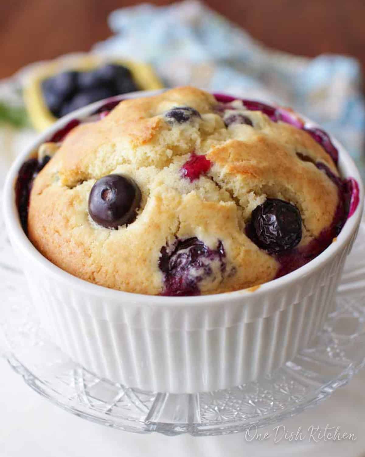 A blueberry muffin in a ramekin plated next to a small bowl of blueberries.