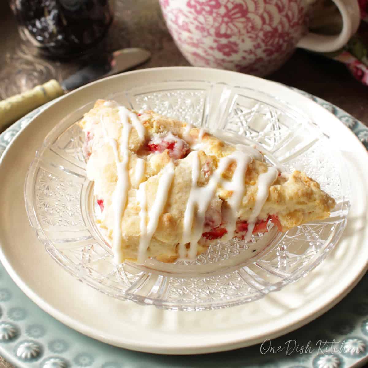 A scone on a plate with drizzled white chocolate next to a cup of coffee and a small jar of jam.