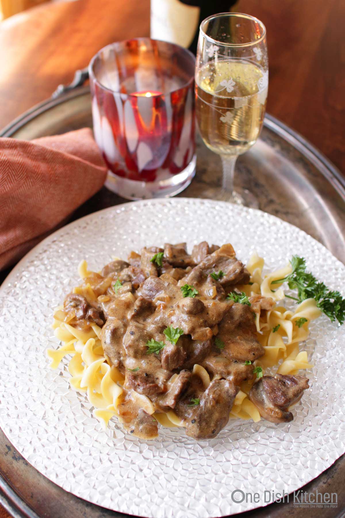 Beef stroganoff plated on a metal tray with a glass of champagne, a red candle, and a red cloth napkin.