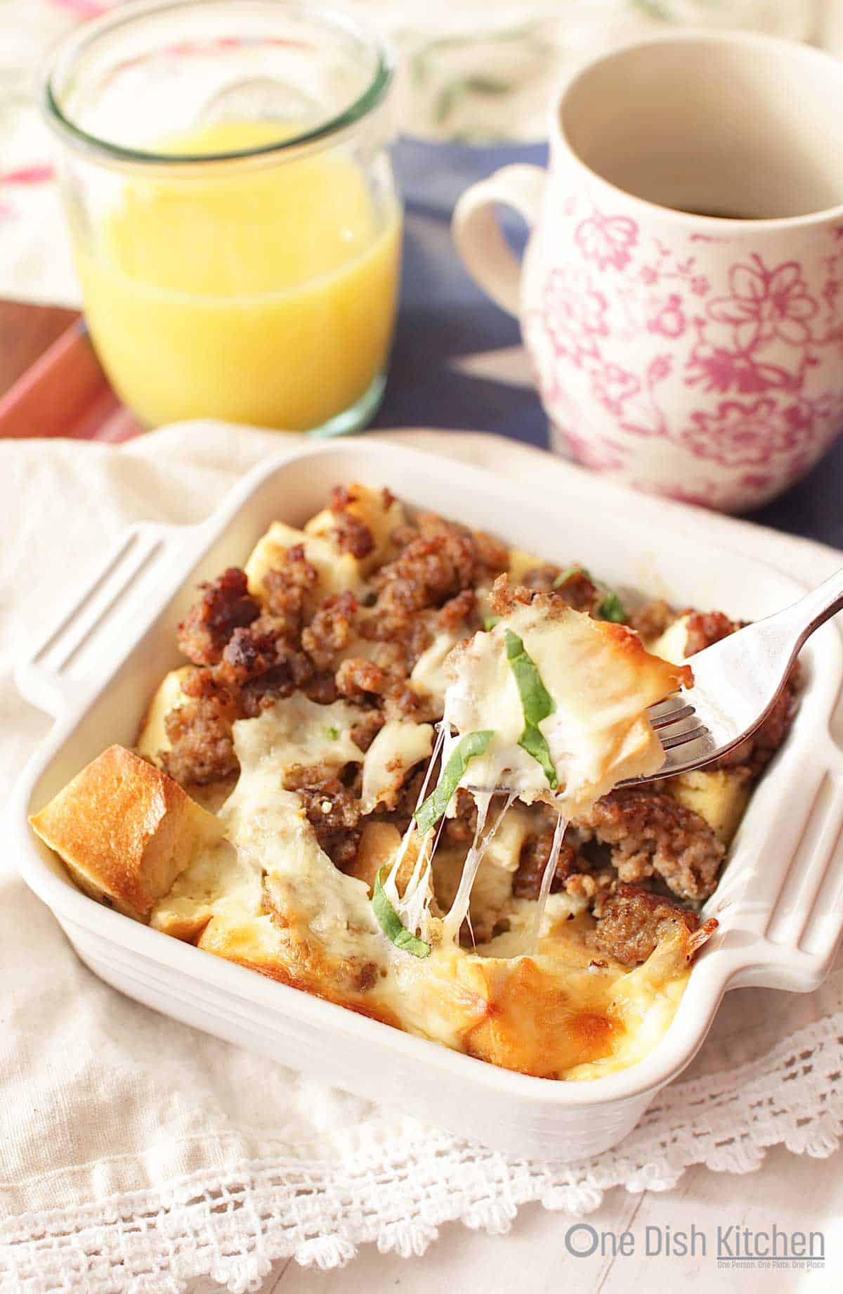 A forkful of breakfast casserole from a small baking dish next to a glass of orange juice and a coffee mug.