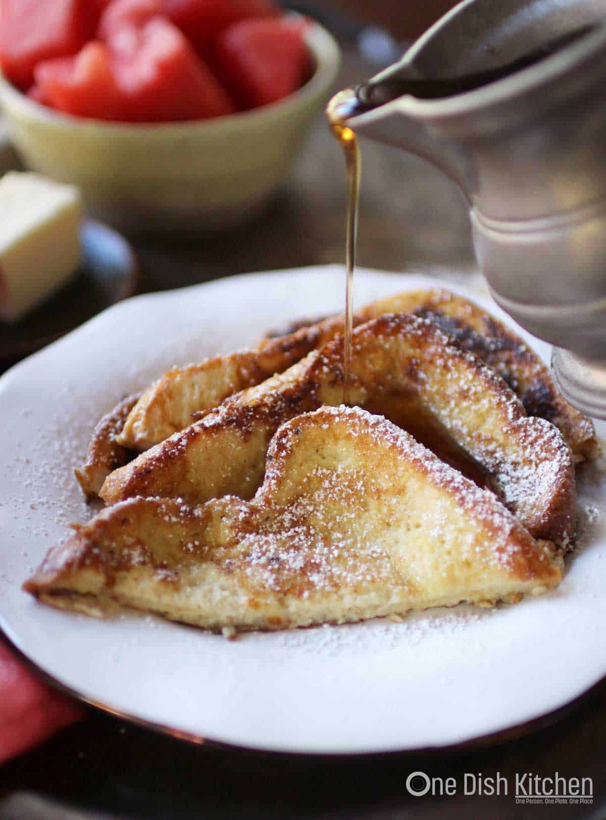 A plate of three slices of french toast dusted with powdered sugar being drizzled with maple syrup
