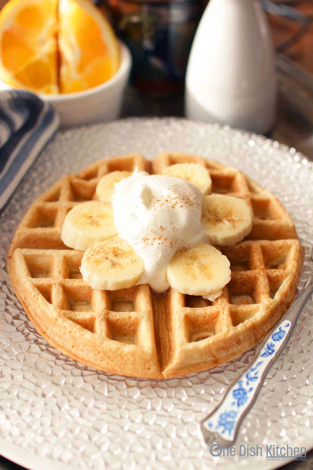 A waffle topped with whipped cream, banana slices, and dusted with cinnamon on a large plate with a fork.