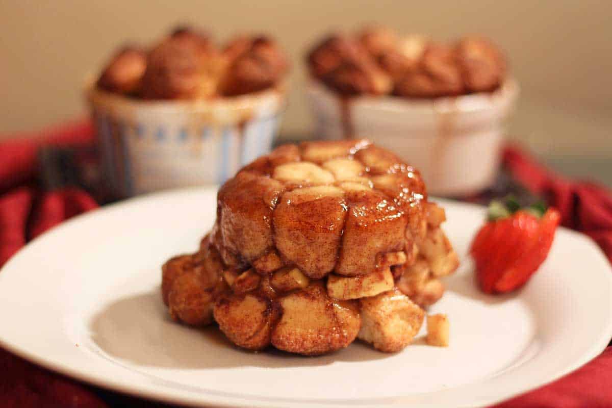 Apple fritter monkey bread on a big plate with a sliced strawberry and two ramekins of mini apple fritter monkey bread in the background