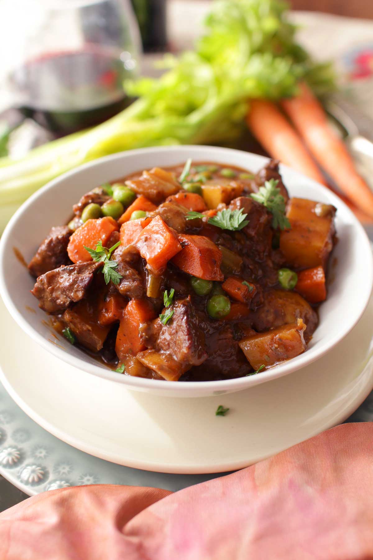 Beef stew in a bowl on a metal tray next to two carrots, a stalk of celery, and a glass of red wine.