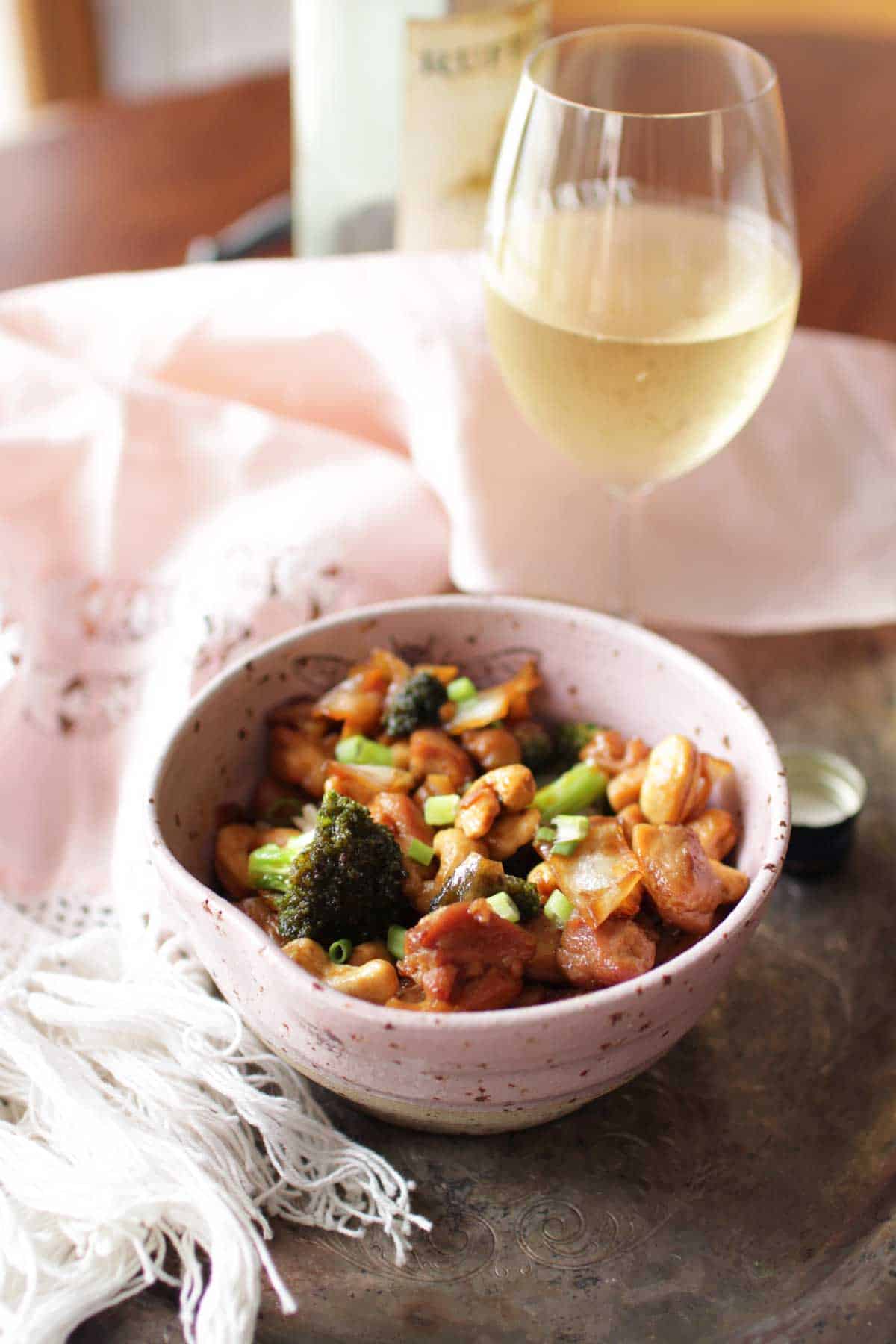 A bowl of cashew chicken next to a glass of white wine on a metal tray