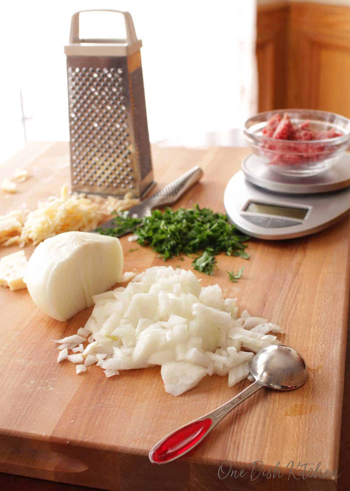 Ingredients for making a meatball on a wooden cutting board- chopped onions, chopped parsley, grated parmesan cheese, and ground beef on a scale.