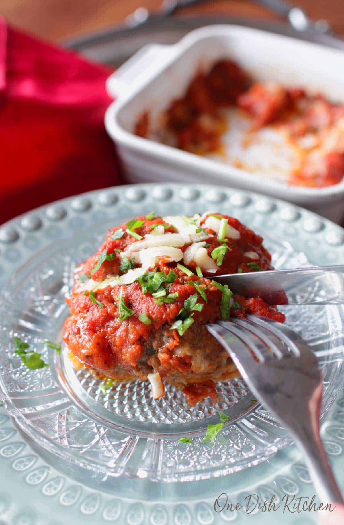 A fork and knife cutting into a meatball topped with tomato sauce and melted parmesan cheese.