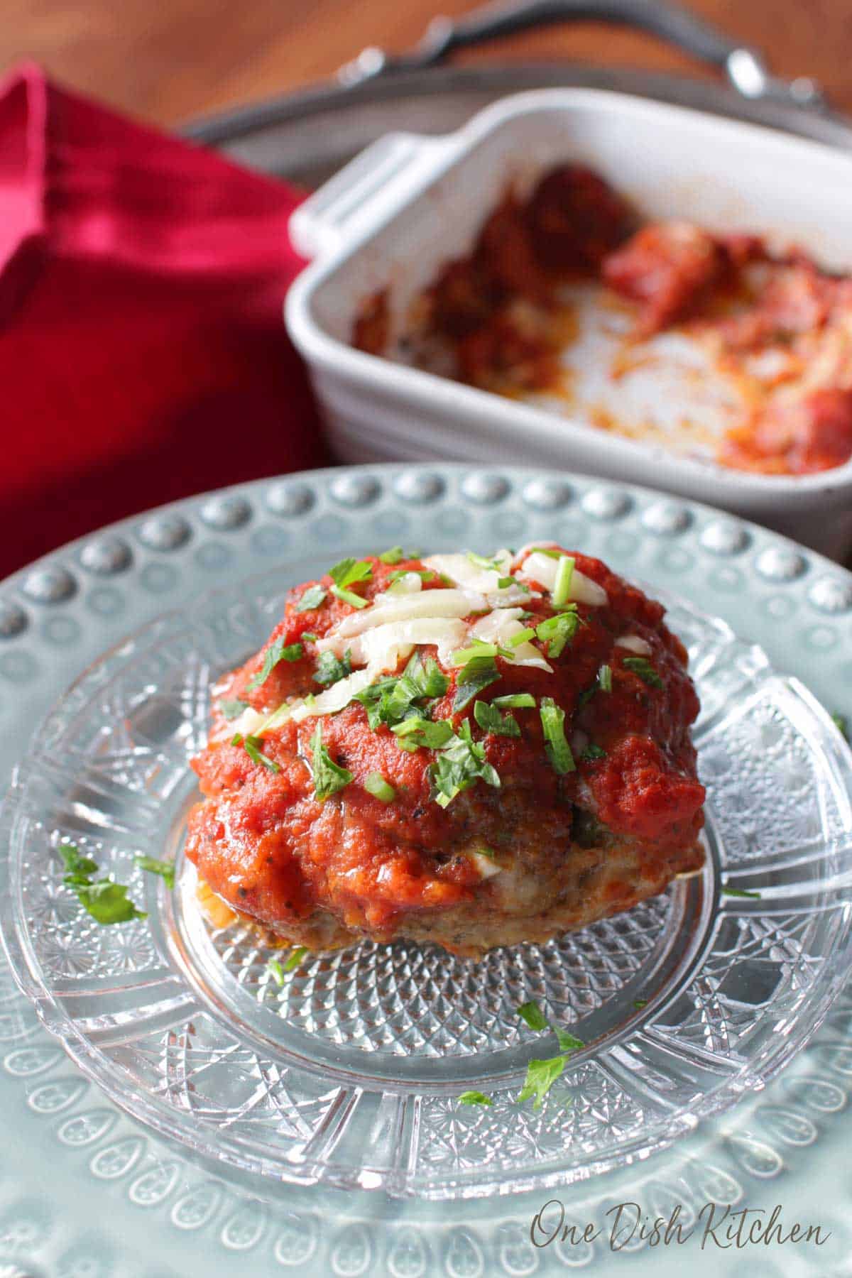 One Italian meatball on a plate topped with tomato sauce and melted parmesan cheese with a baking dish in the background.