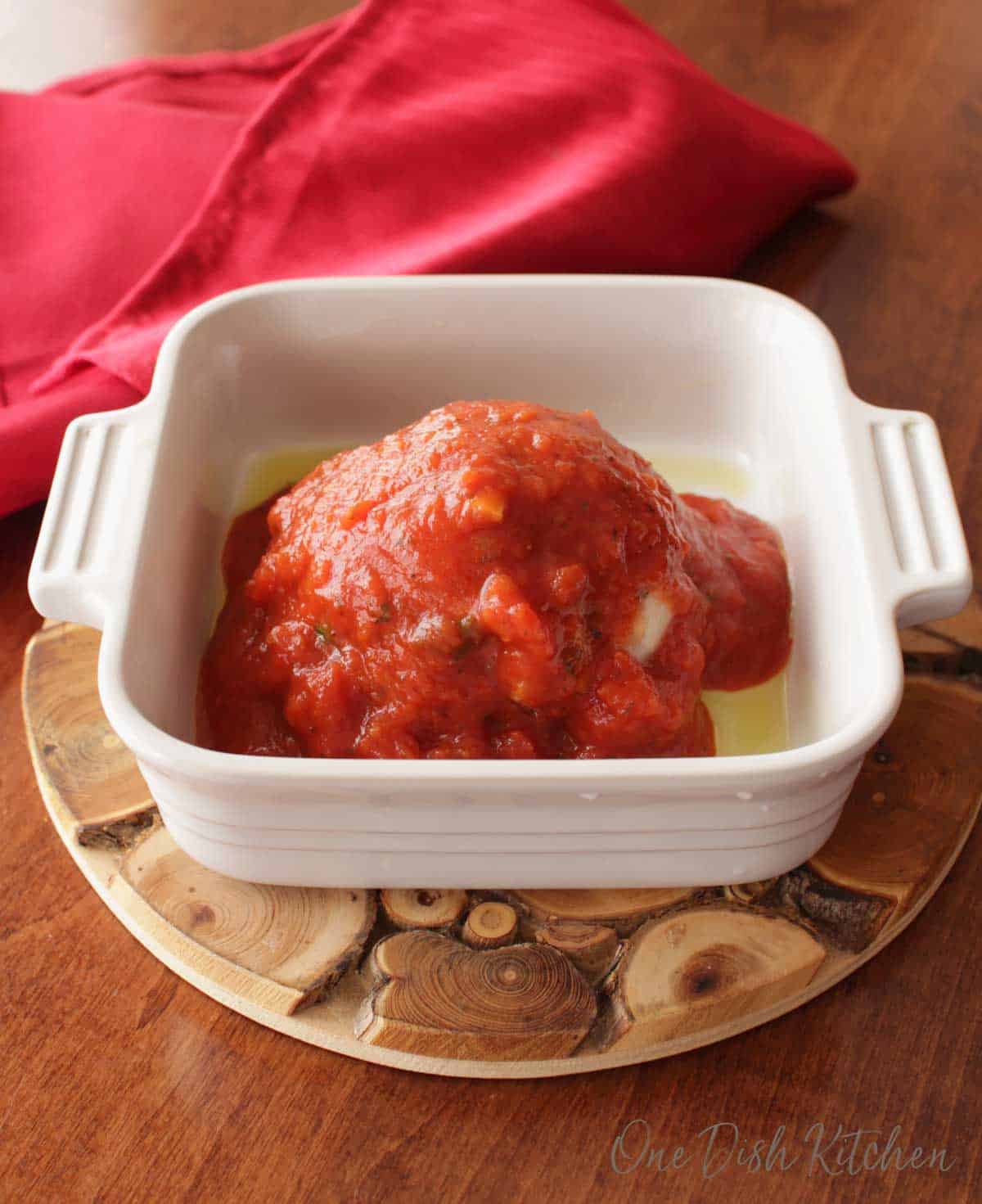A meatball topped with tomato sauce in a small baking dish on a wooden trivet. 