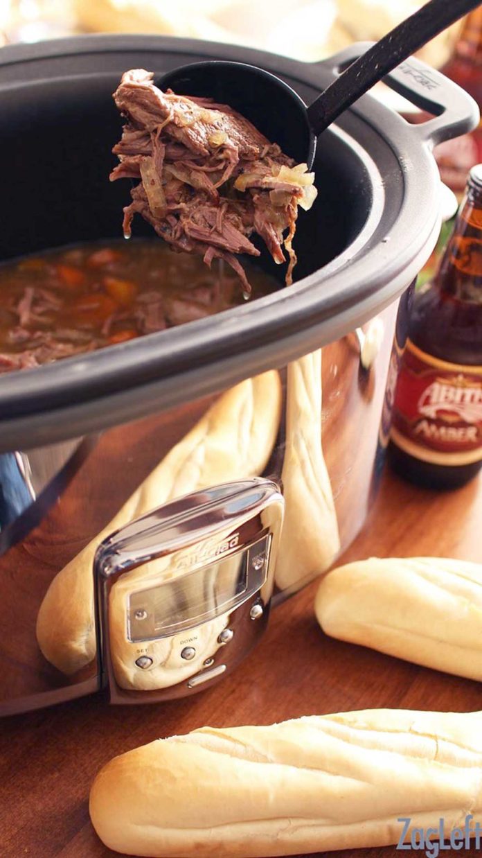 A spoonful of pot roast from a slow cooker next two a bottle of beer and two baguettes.