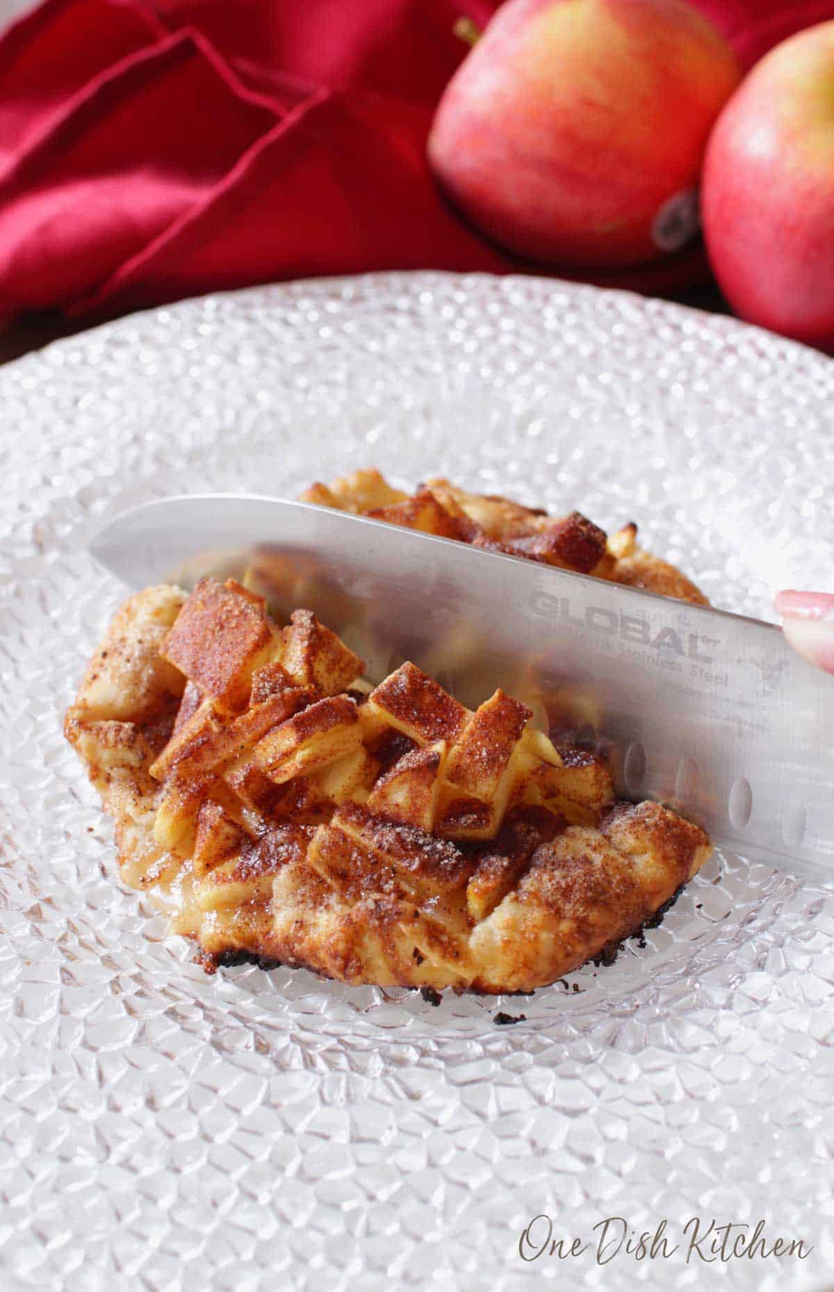 A knife cutting into an apple galette topped with cinnamon and sugar plated with apples and a red cloth napkin in the background.