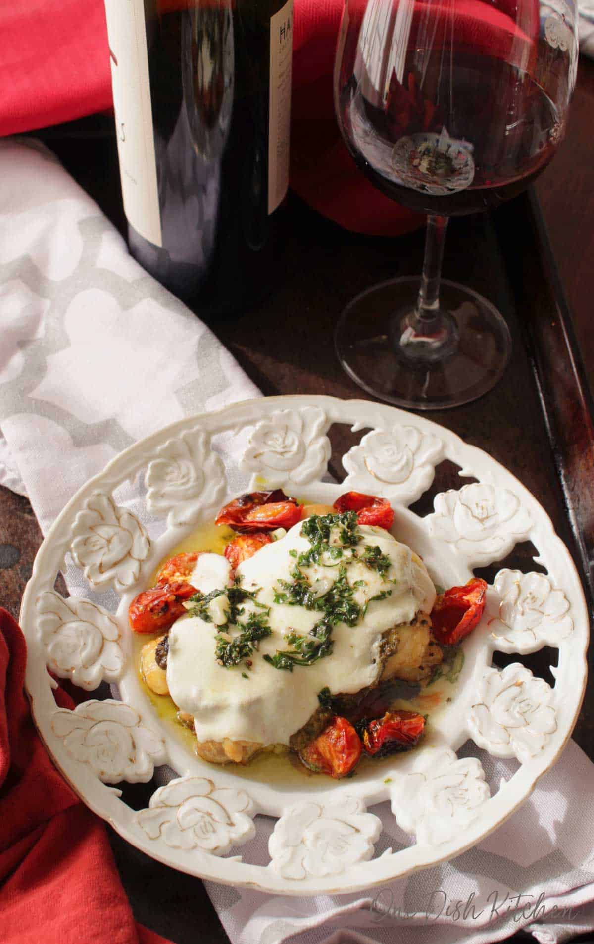 An overhead view of Chicken margherita on a plate with roasted cherry tomatoes next to a glass and bottle of red wine