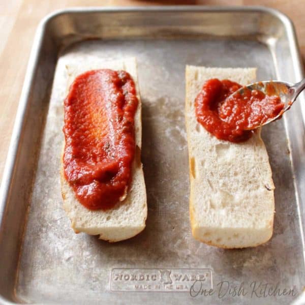 spreading tomato sauce on french bread for a french bread pizza | one dish kitchen