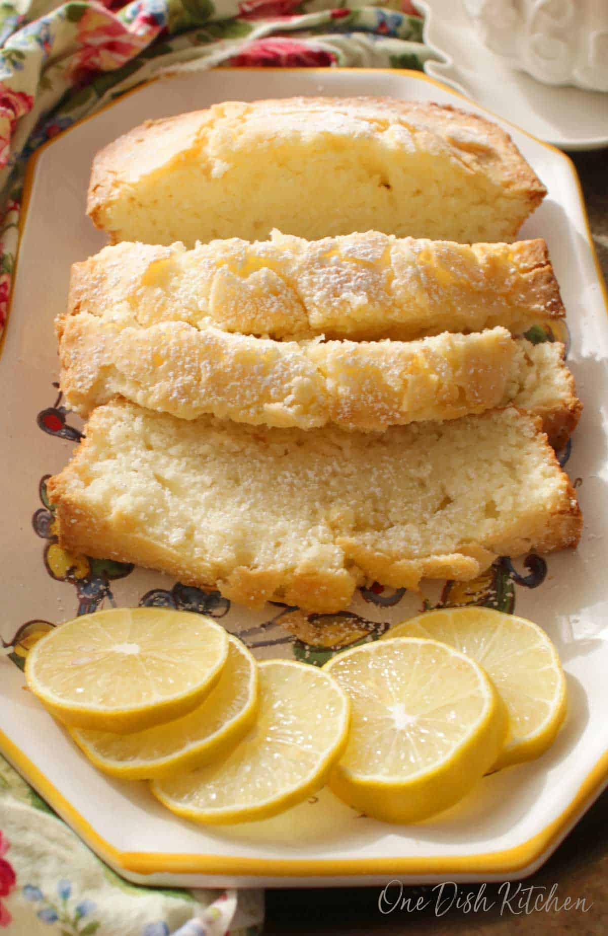 Four slices of pound cake dusted with powdered sugar on a plate with five decorative lemon wheels.