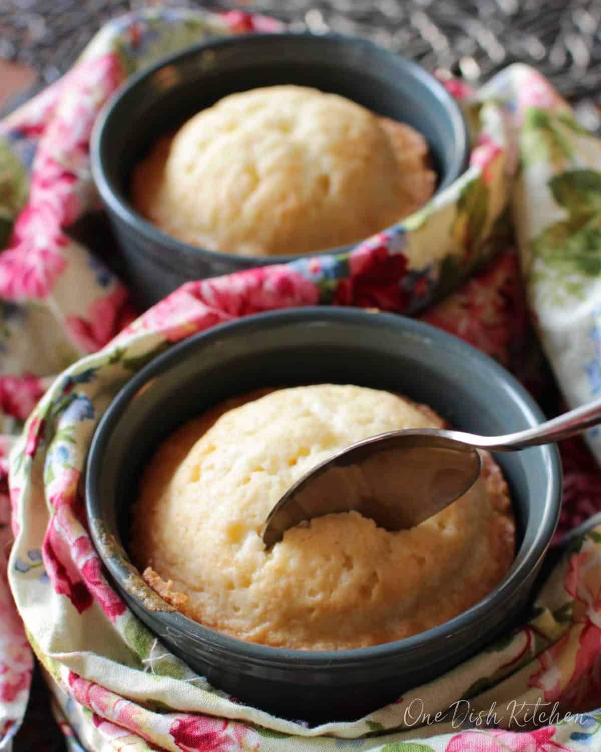 A spoon in a pound cake baked in a ramekin next to another pound cake in a ramekin all wrapped in a floral cloth napkin.