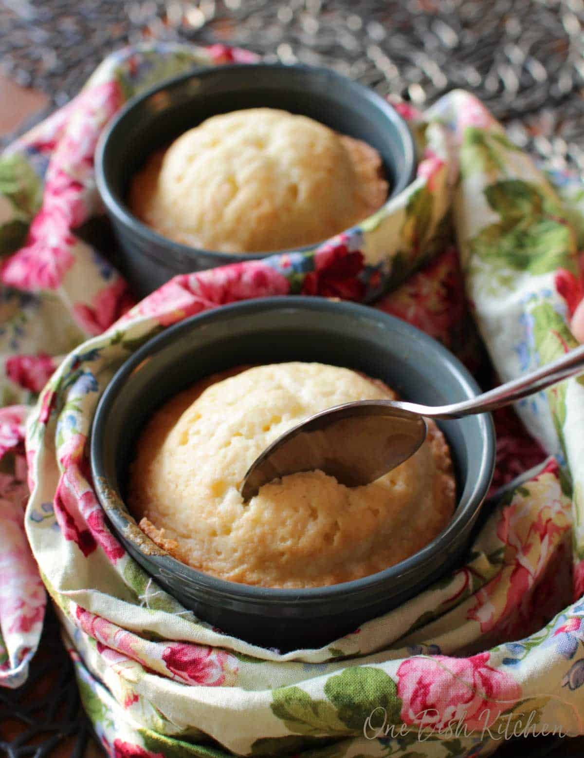 A spoon in a pound cake baked in a ramekin next to another pound cake in a ramekin all wrapped in a floral cloth napkin