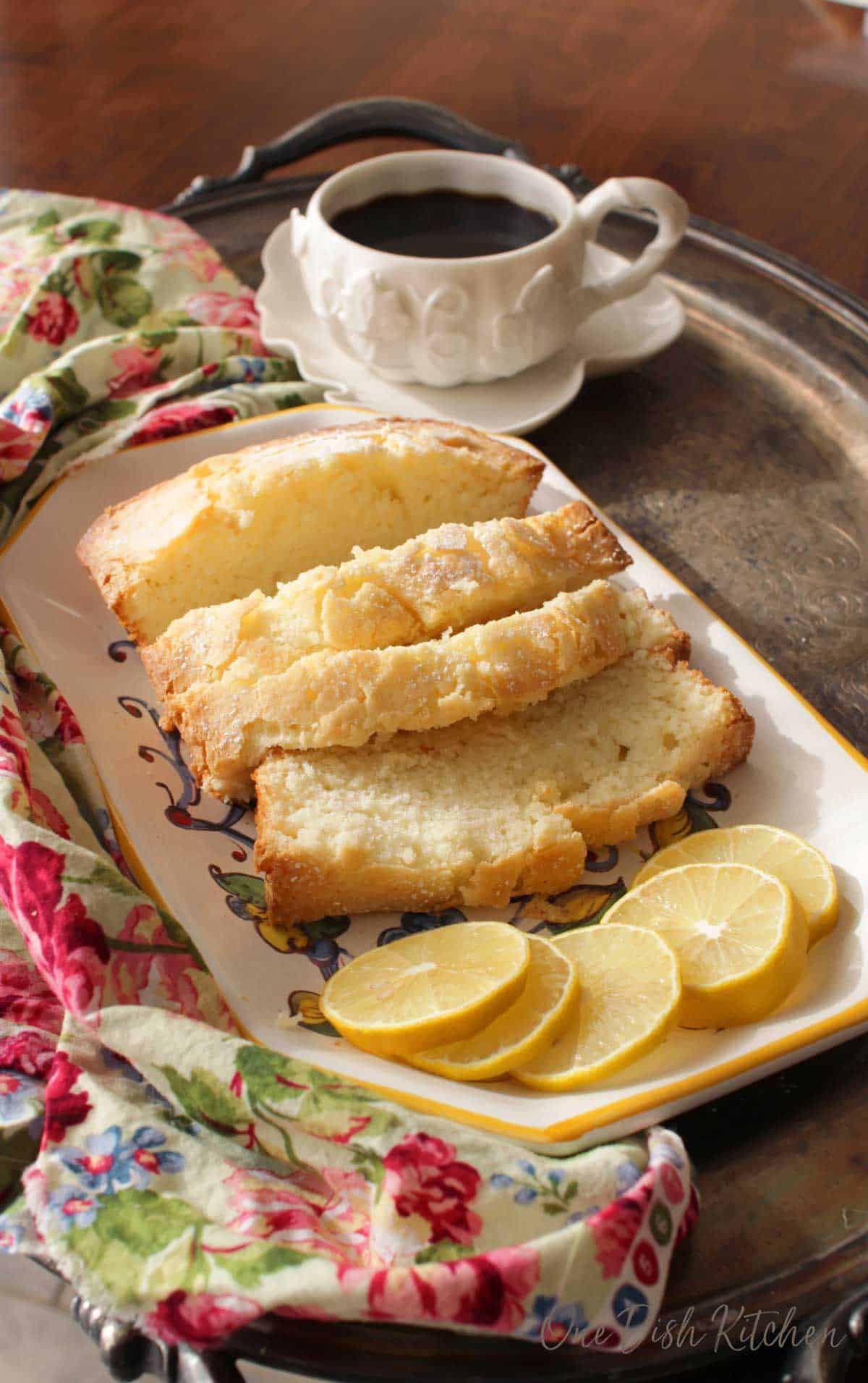 Four slices of pound cake dusted with powdered sugar on a plate with five decorative lemon wheels on a metal tray with a mug of coffee and a floral cloth napkin