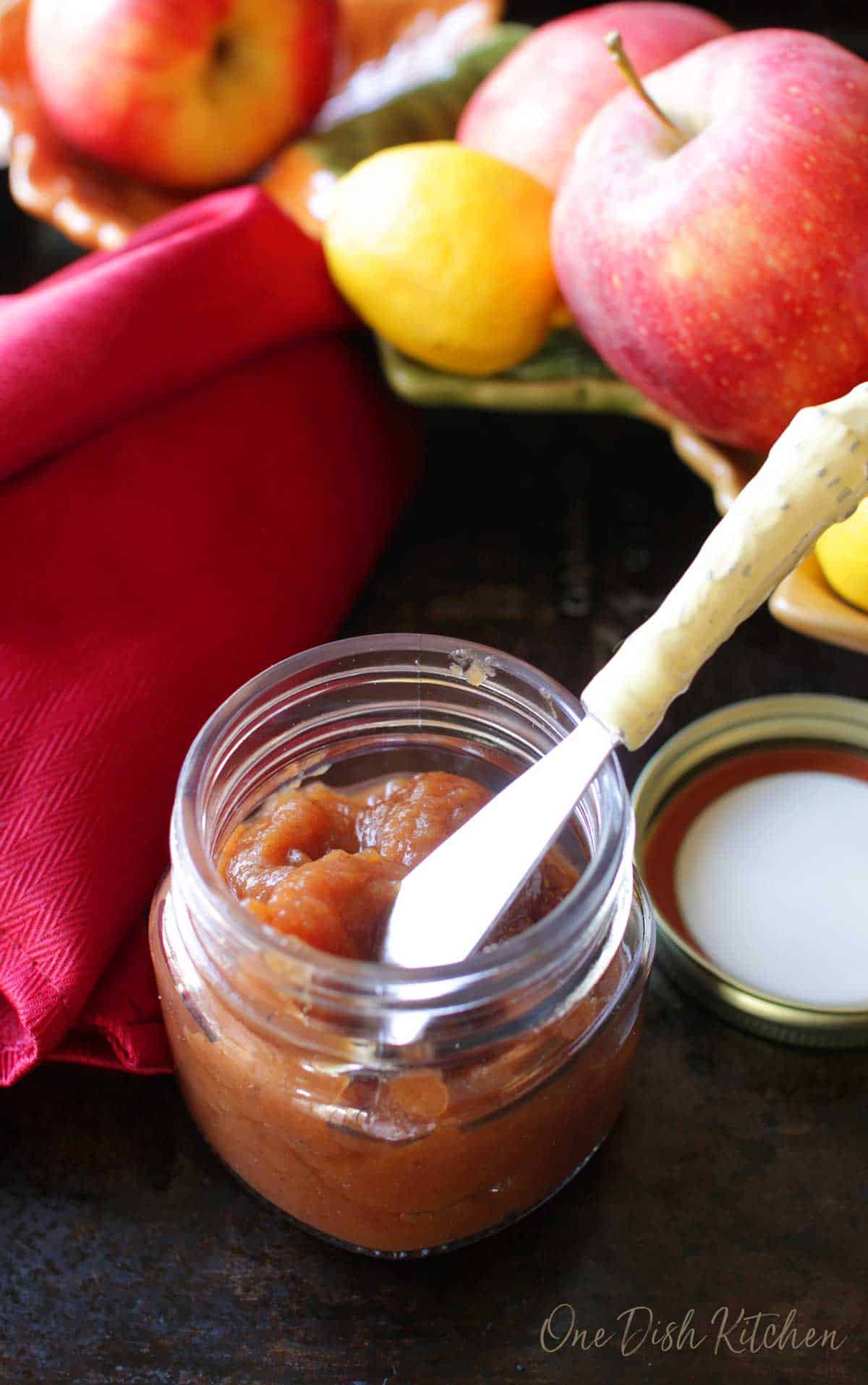 Apple butter in a small jar with a knife on a dark surface with a red cloth napkin and a bowl of apples and lemons.