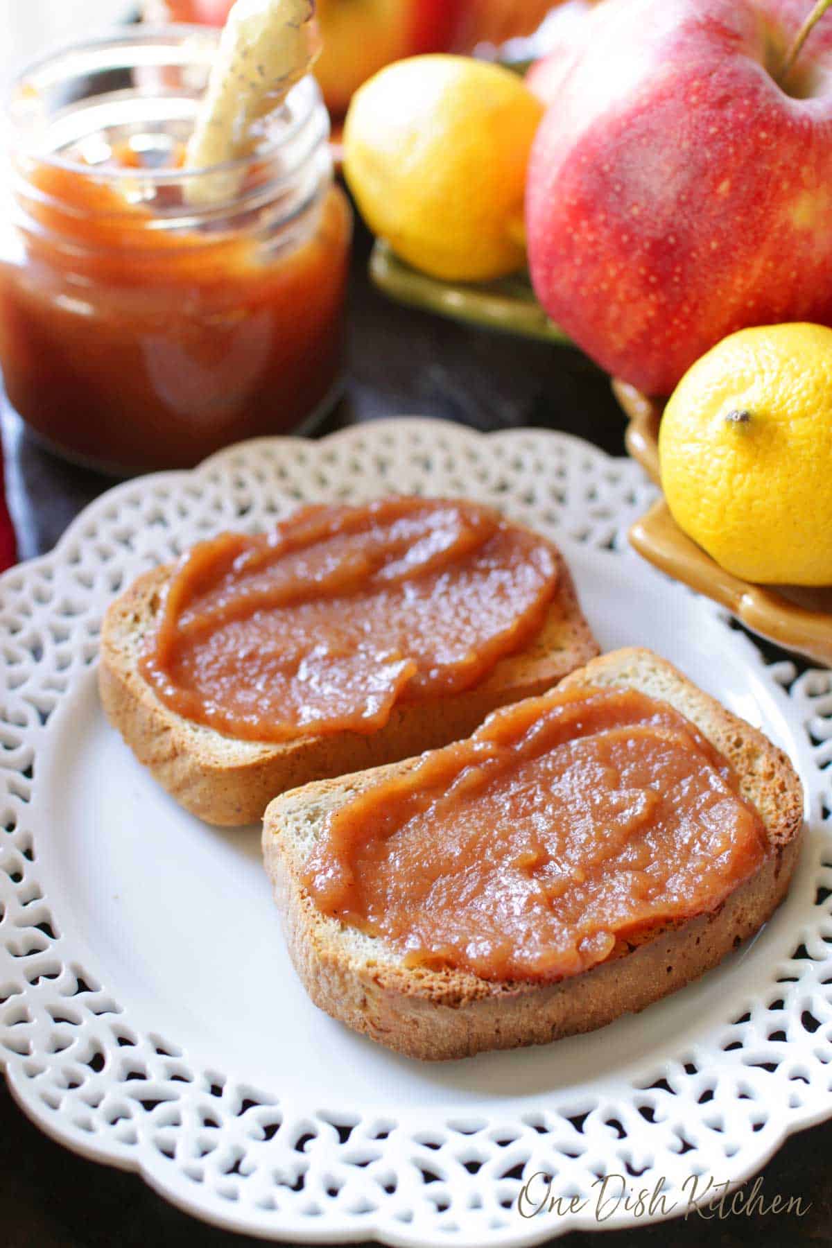 Apple butter spread on two slices of toast plated next to a small jar of apple butter with a knife and a bowl of apples and lemons.