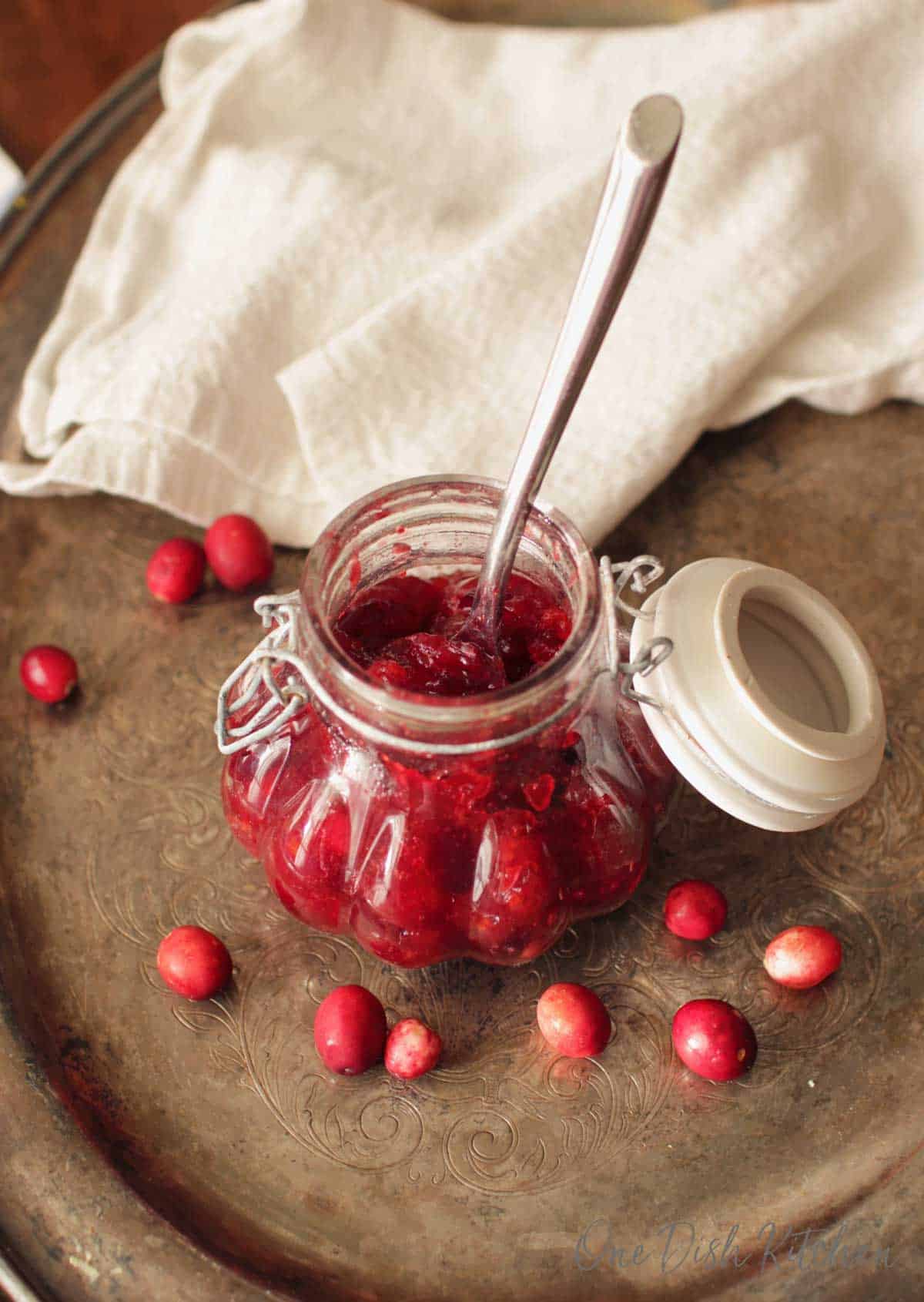An overhead view of a jar of cranberry jam with a spoon on a metal tray with scattered cranberries and a white cloth napkin.