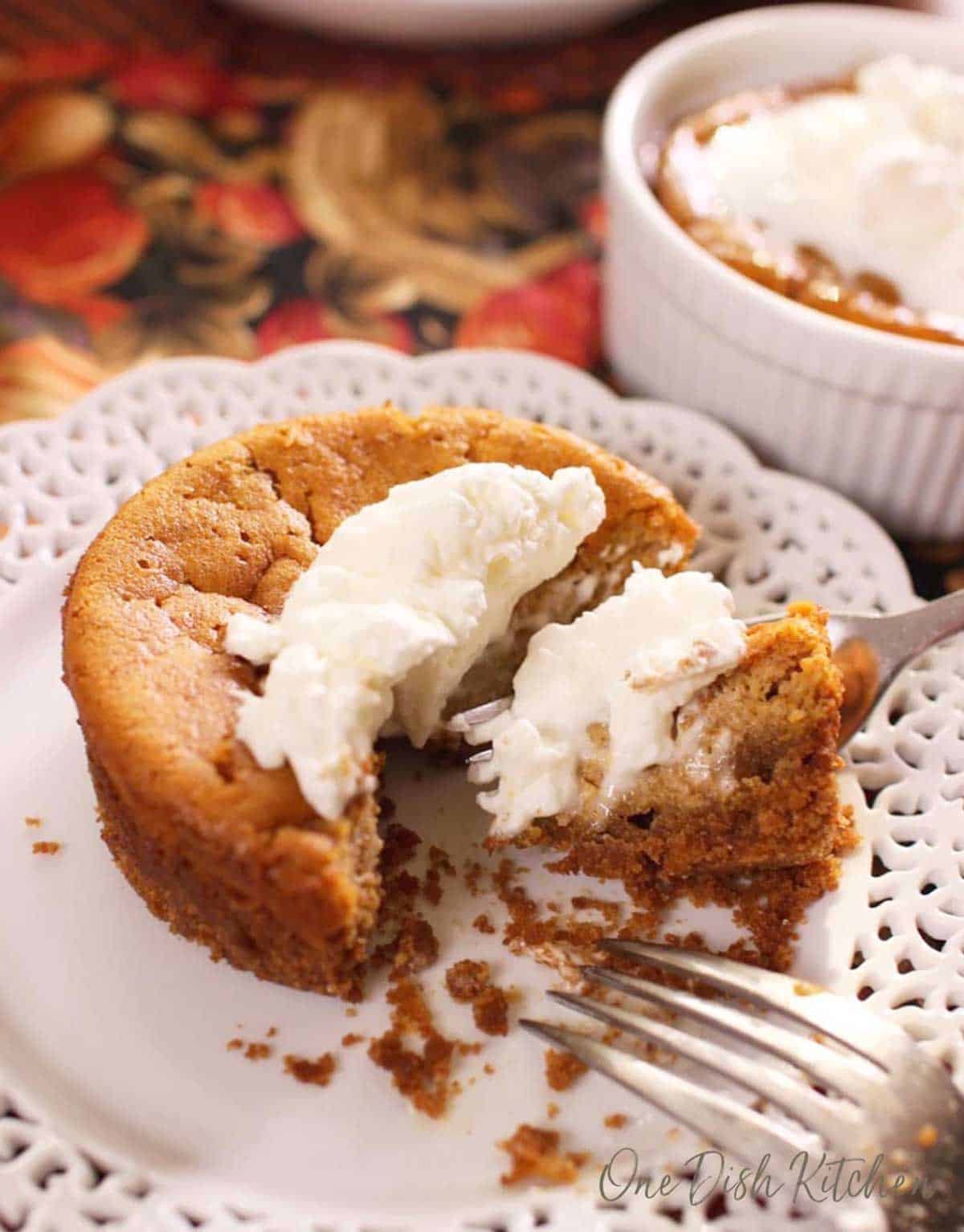 a small round pumpkin pie topped with whipped cream on a white lace plate on top of a orange and gold tablecloth.