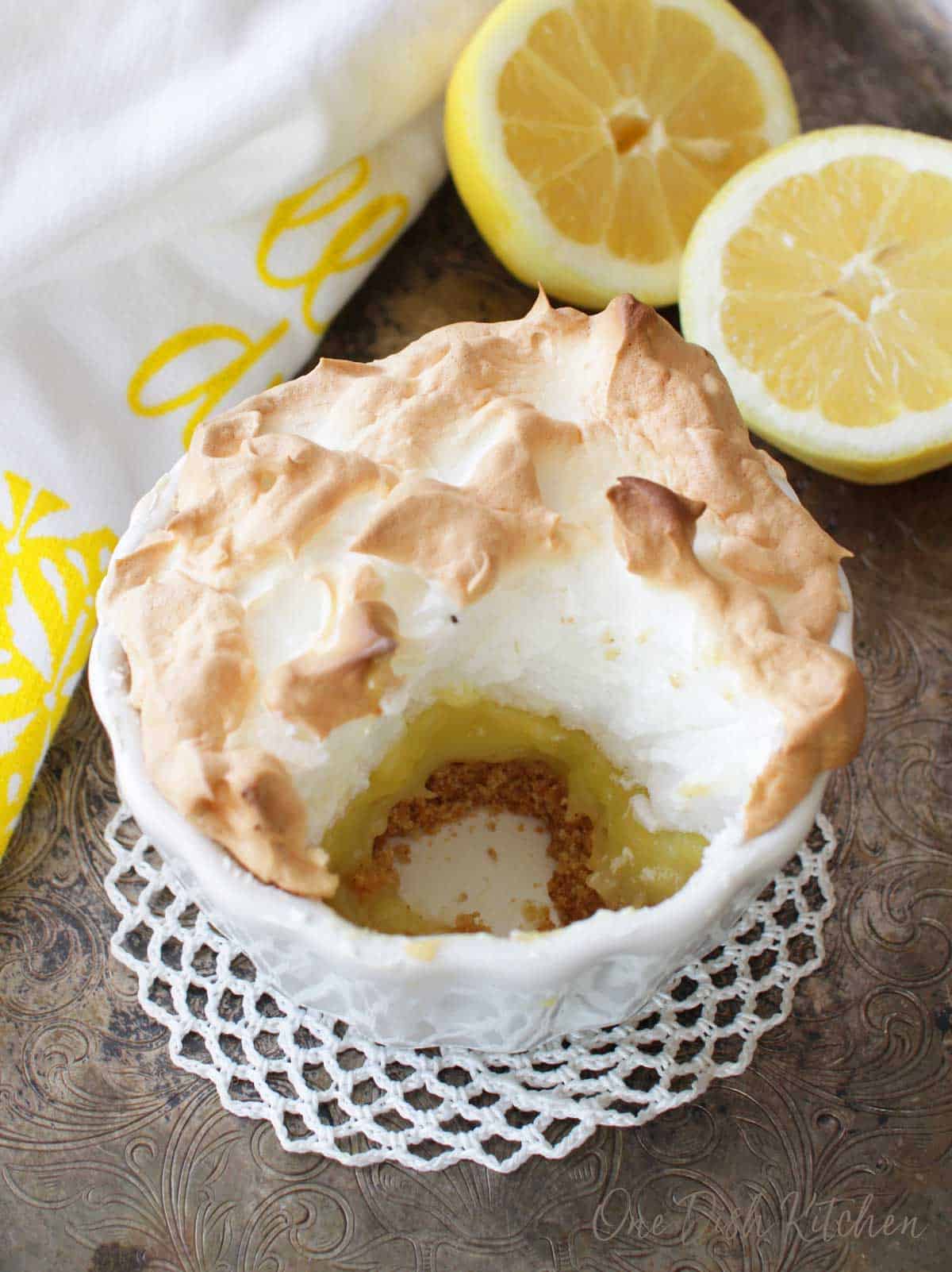 A small round lemon meringue pie with a round section removed next to sliced lemons on a silver tray.