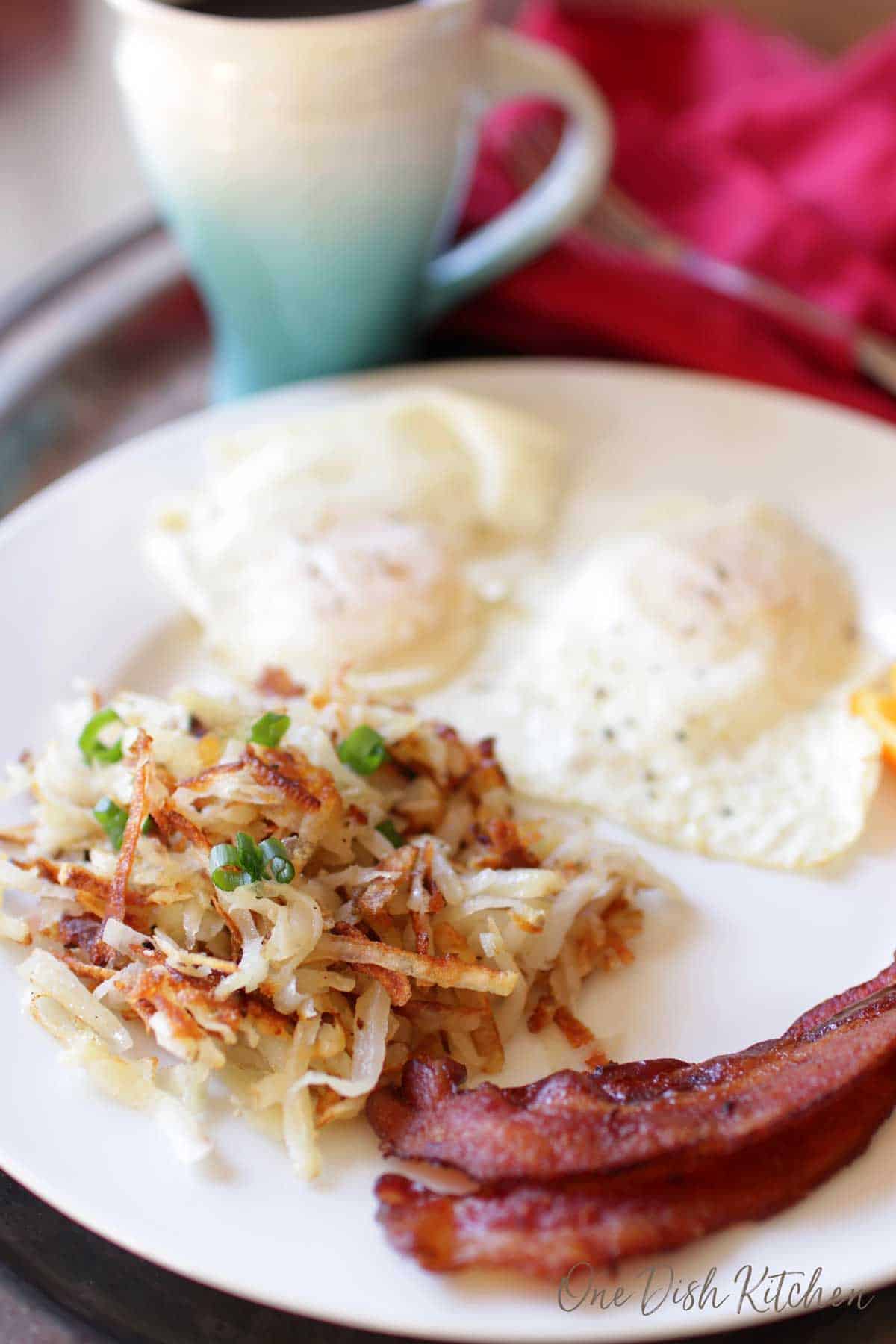 Hash browns with two slices of bacon and two eggs plated on a metal tray with a mug of coffee.