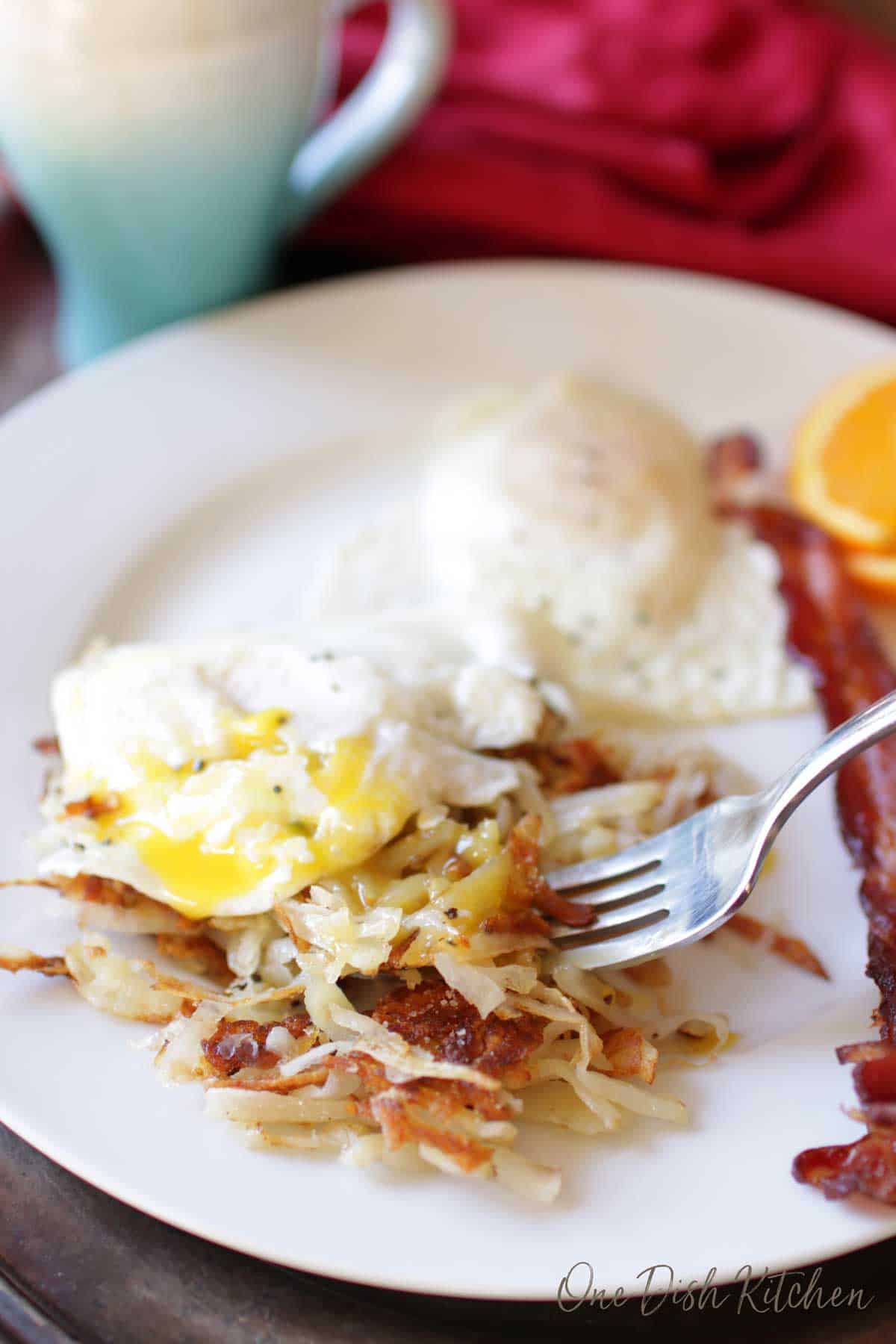 A forkful of hash browns and runny eggs from a plate with slices of bacon and orange slices.