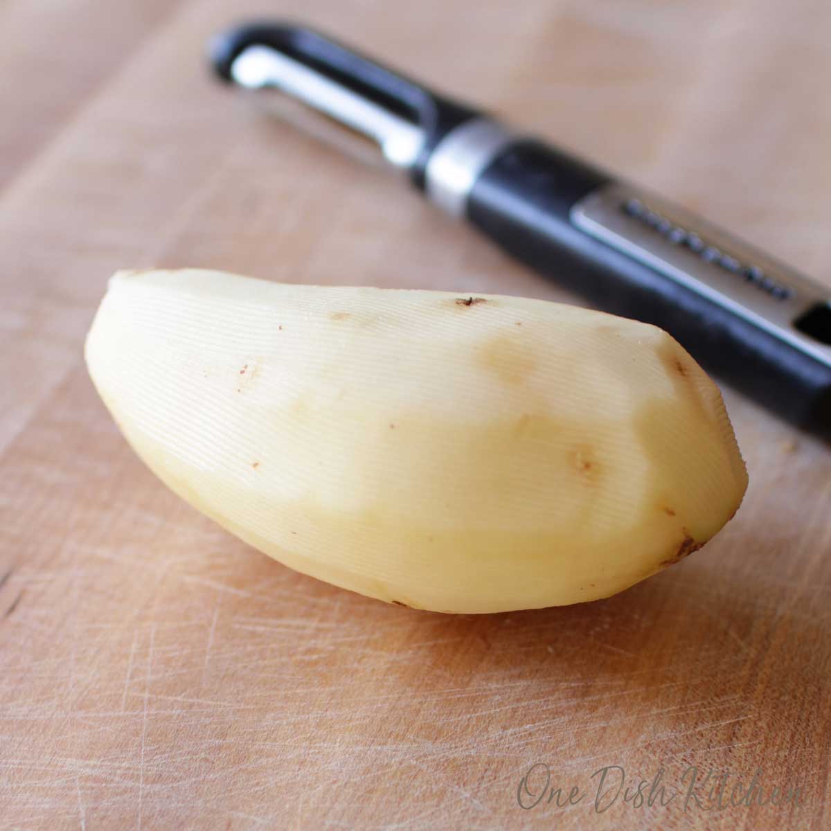 One peeled potato next to a peeler on a wooden cutting board.