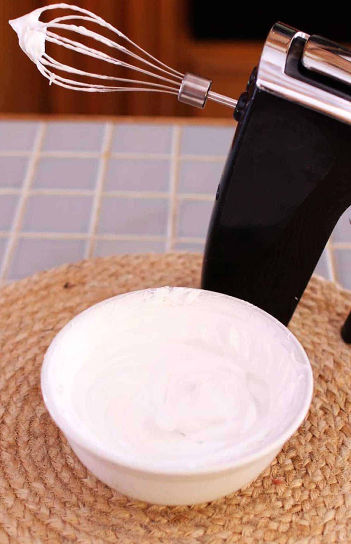 A bowl of freshly whipped cream next to an electric hand mixer with cream on the whisk.