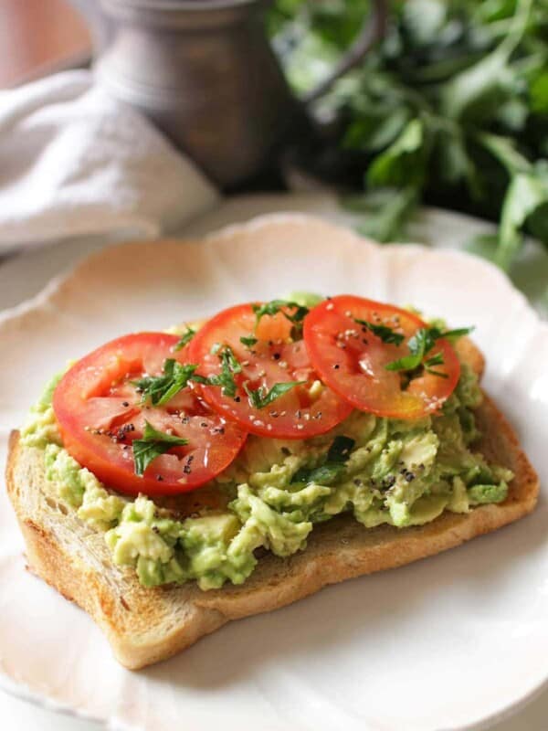 a slice of bread topped with mashed avocados on a white plate next to basil leaves